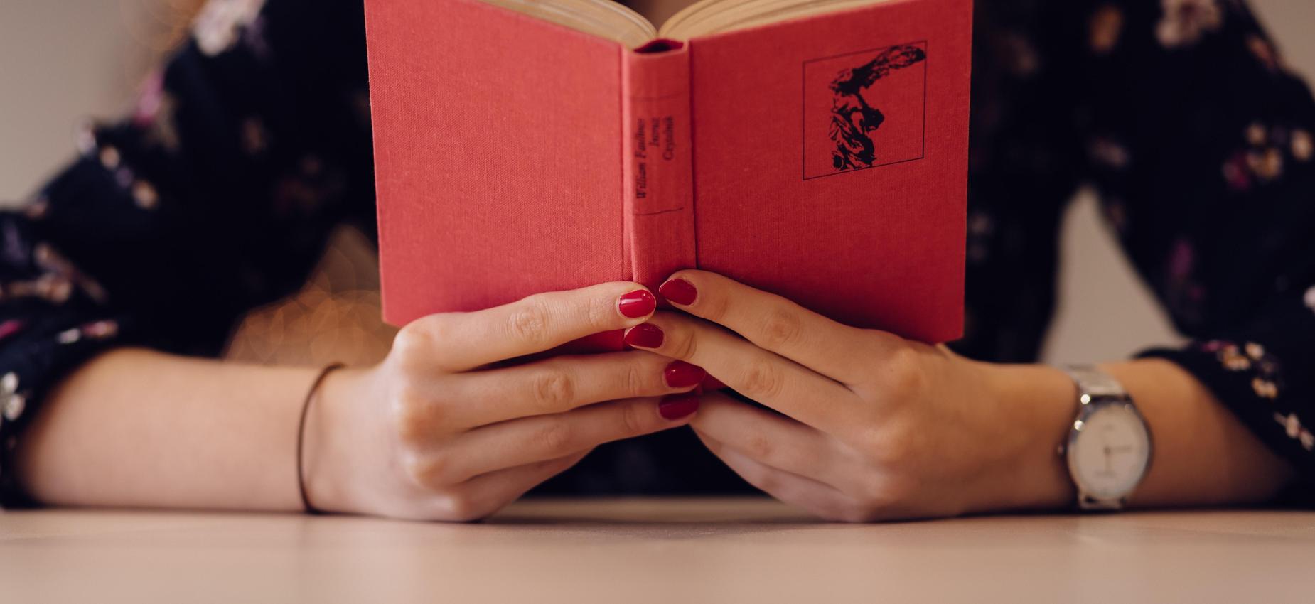 Person reading a red book with floral attire and a watch on wrist.