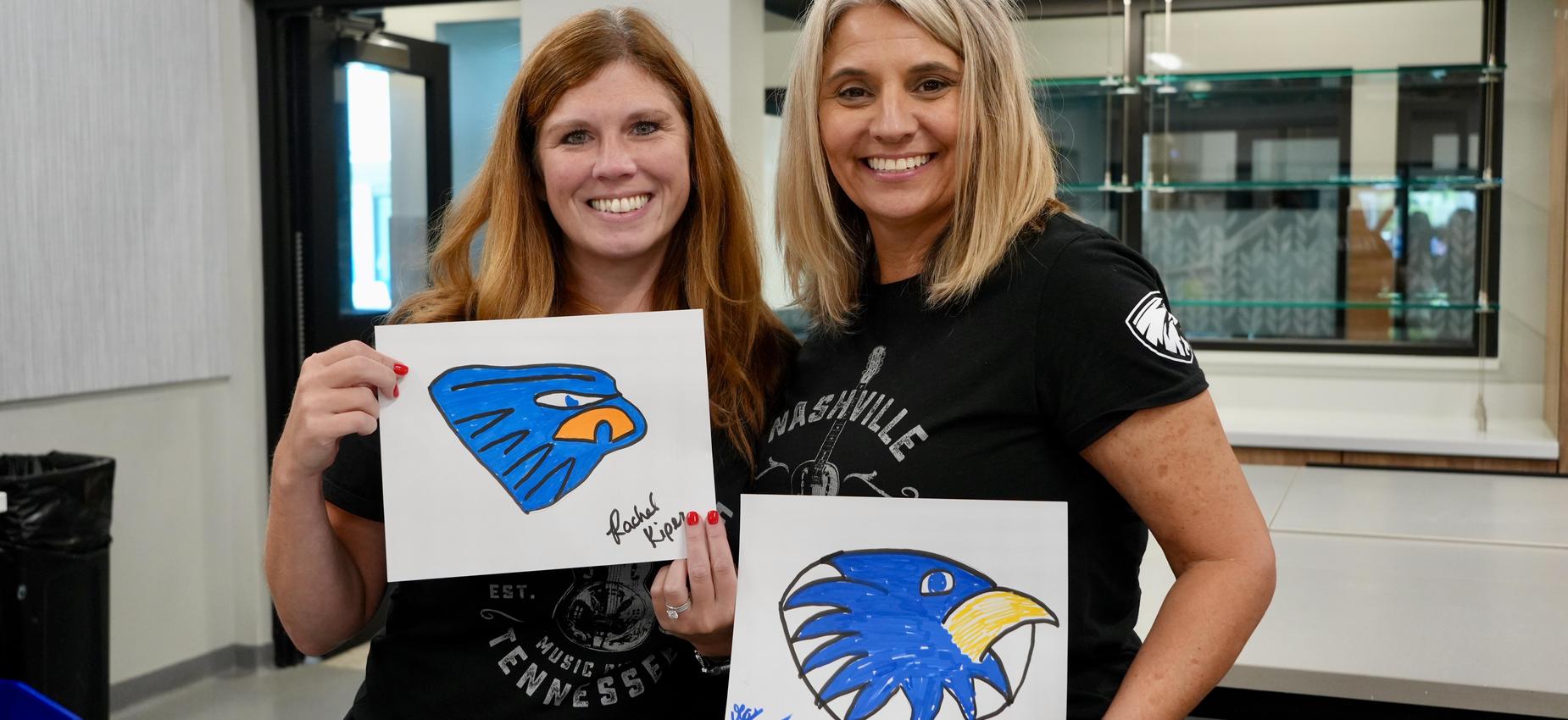 Two women holding drawings of birds, smiling in a classroom setting.