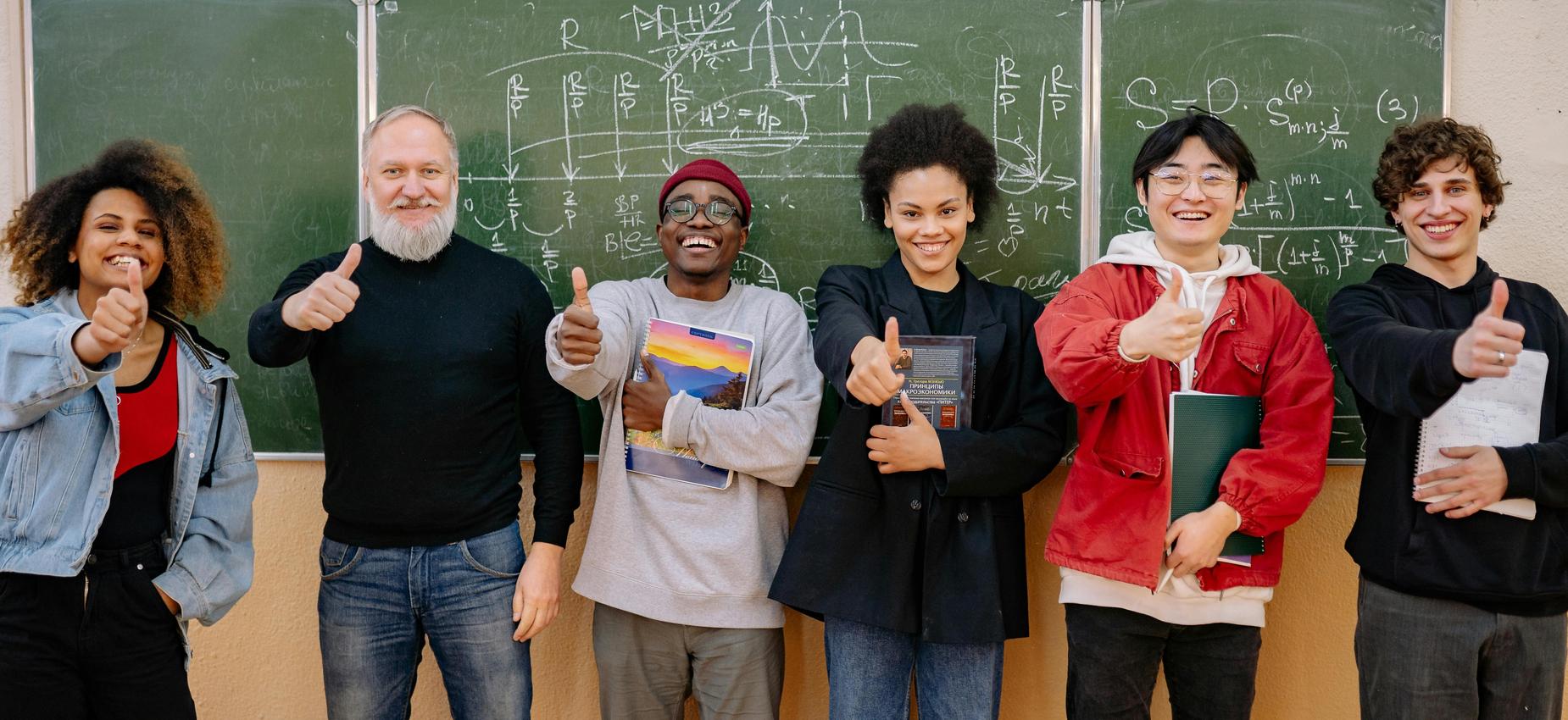 Group of diverse students giving thumbs up in front of a chalkboard.