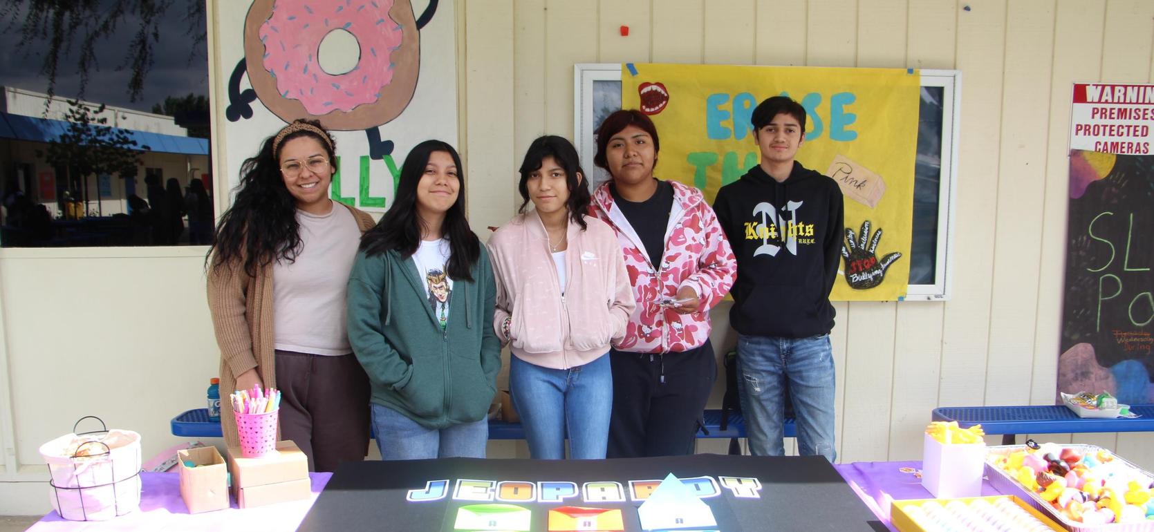 Five students pose together at a Jeopardy-themed event with colorful game boards.