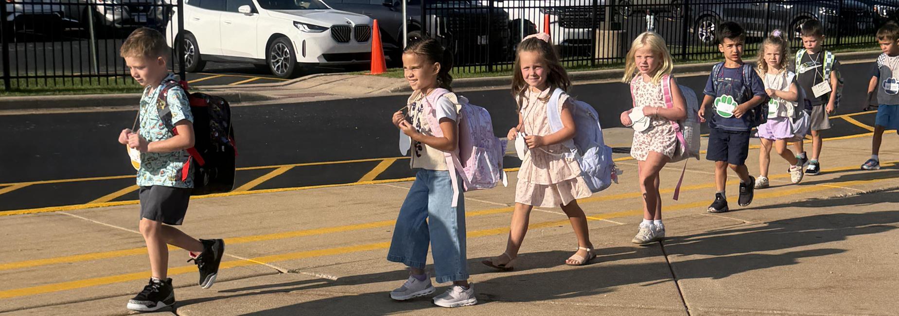 Children walking along a sidewalk after school, wearing casual clothing and backpacks.