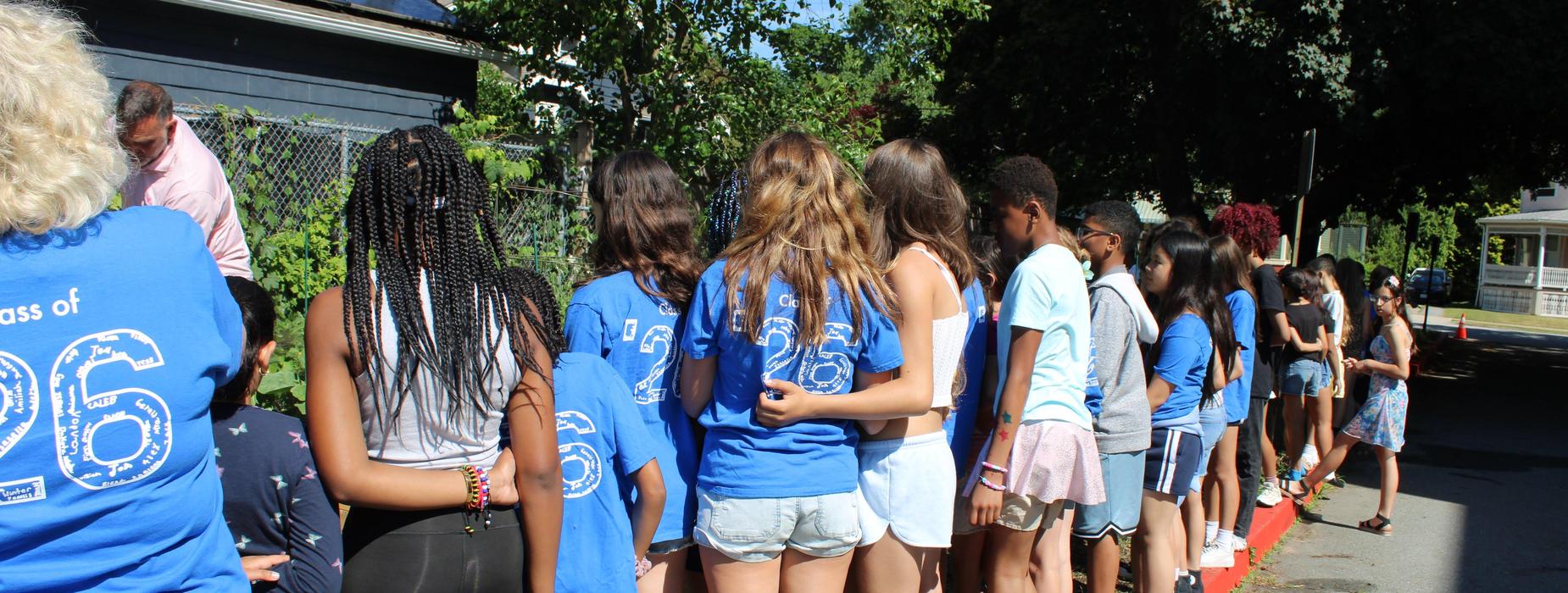 Students and the principal stand in a line along the curb, watching the time capsule be buried. Their blue shirts are the Class of 2026 shirts.