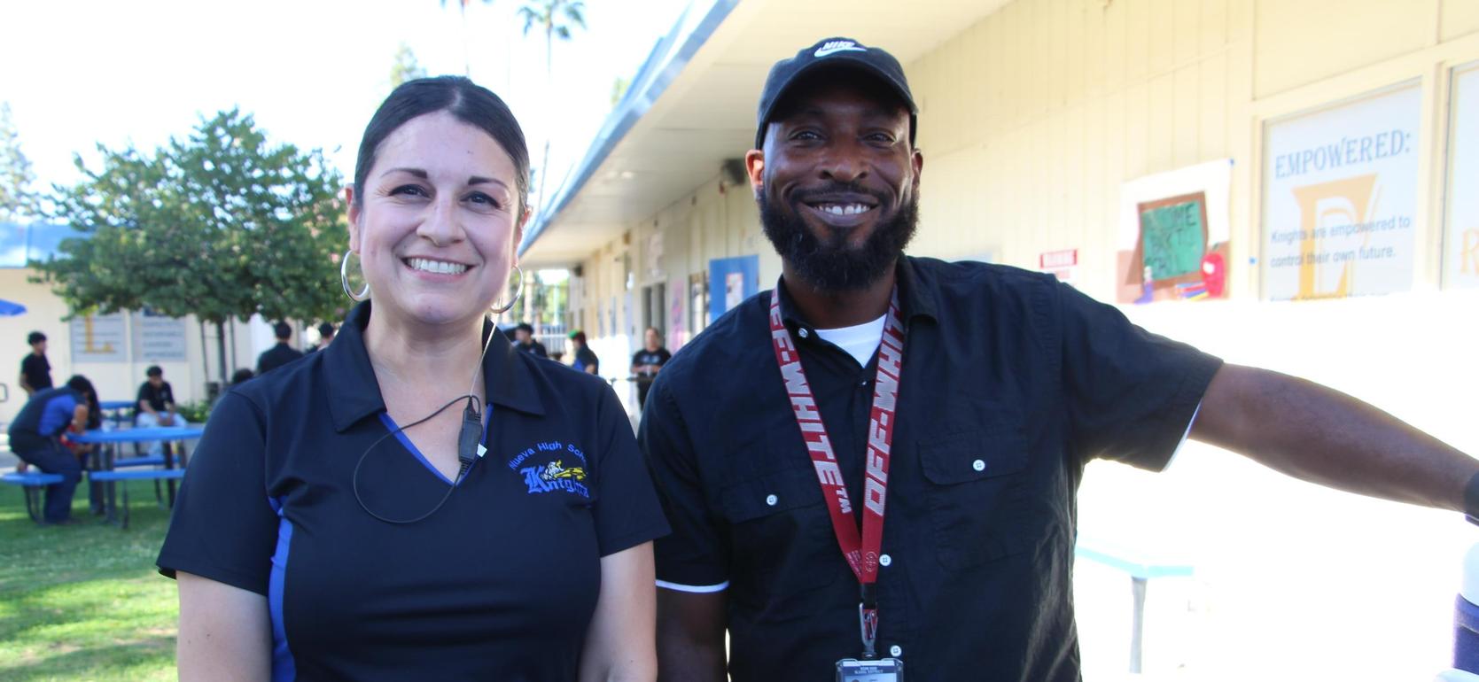 Two smiling individuals standing outdoors near a school building.