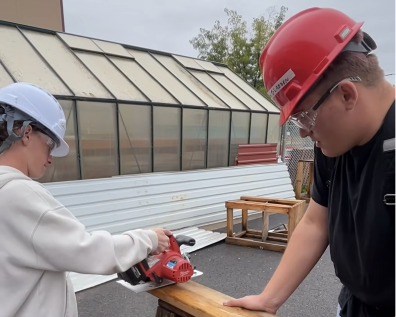 A high school student uses a circular saw while another student secures the board.