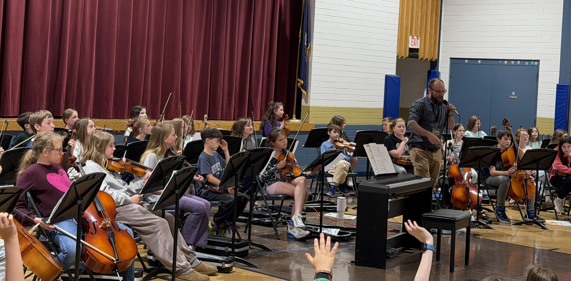 Young orchestra musicians practicing together under a basketball hoop indoors.