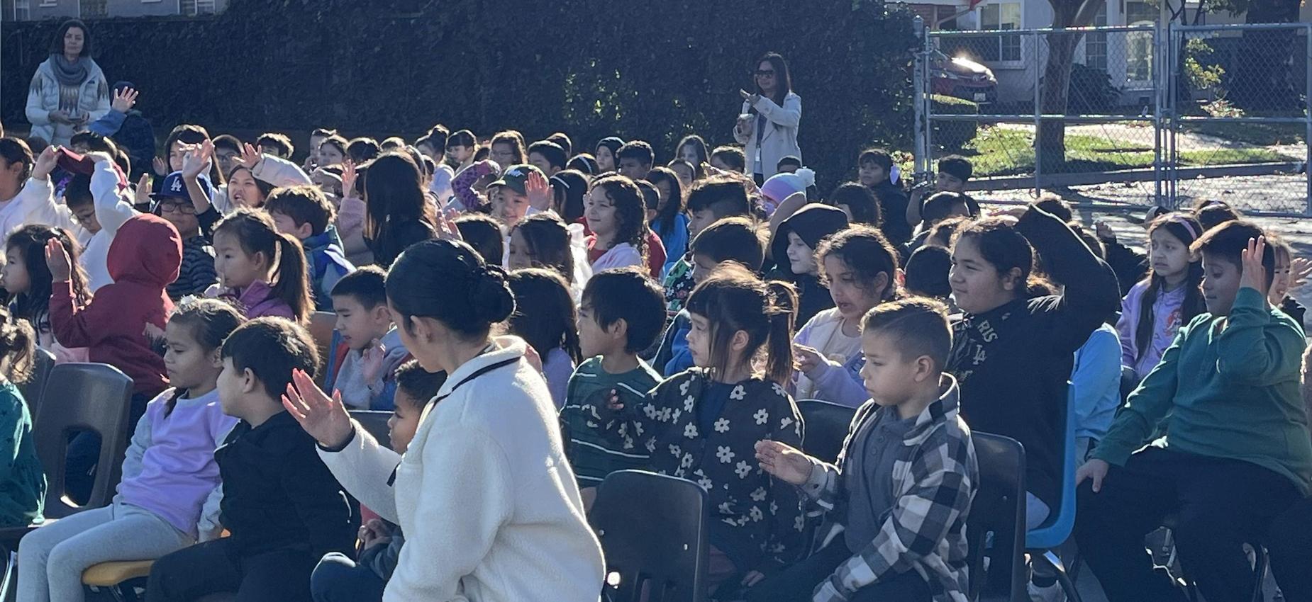 A diverse group of children and adults seated outdoors, some raising hands in a crowd.