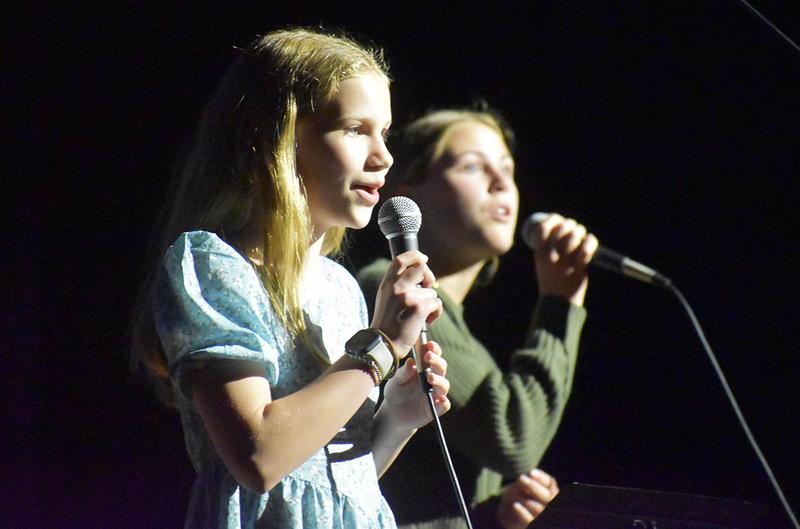 Two girls singing into microphones, one in a blue dress and the other in a sweater.