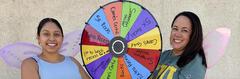 Two women with butterfly wings beside a colorful prize wheel.