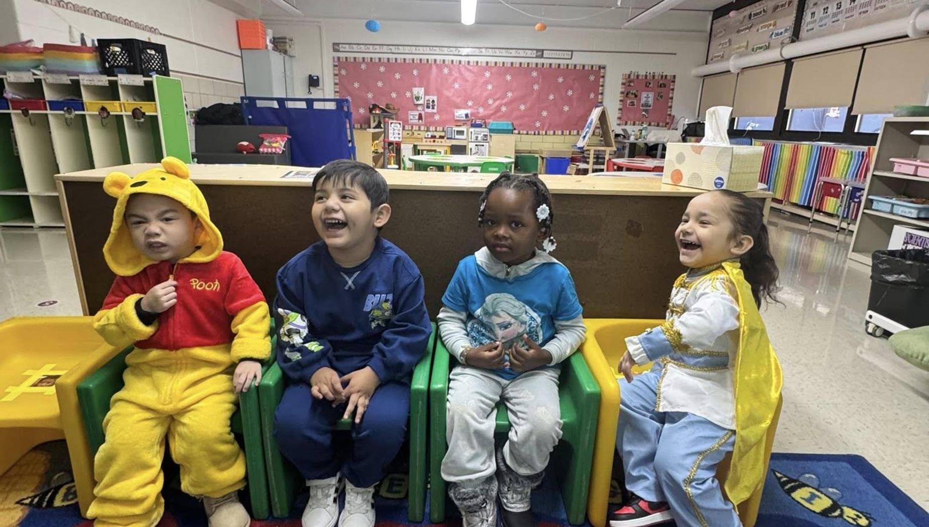 Four children laughing while sitting on colorful chairs in a cheerful classroom.