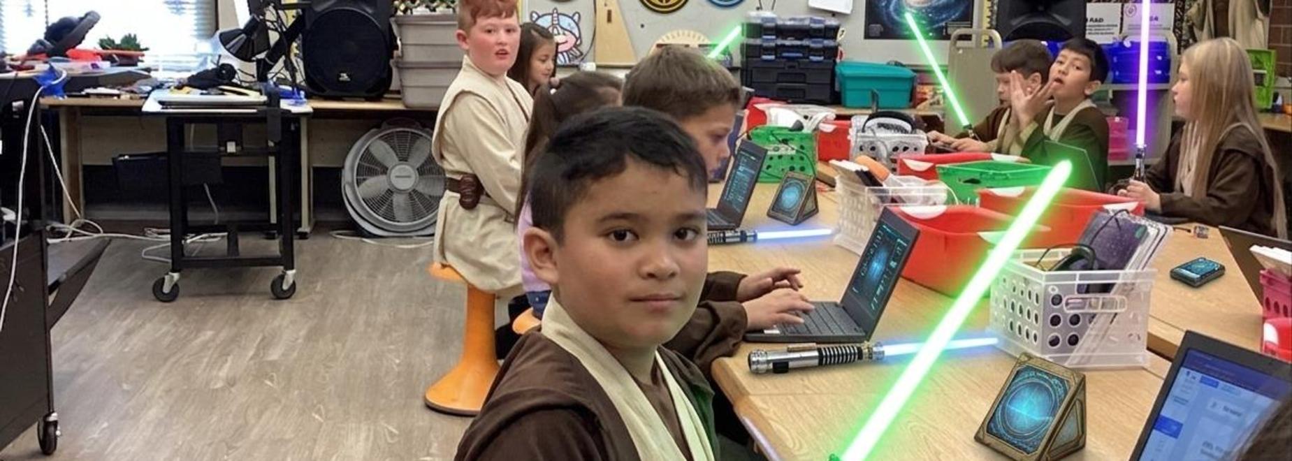 A child dressed as a Jedi holds a lightsaber while classmates work at tables.