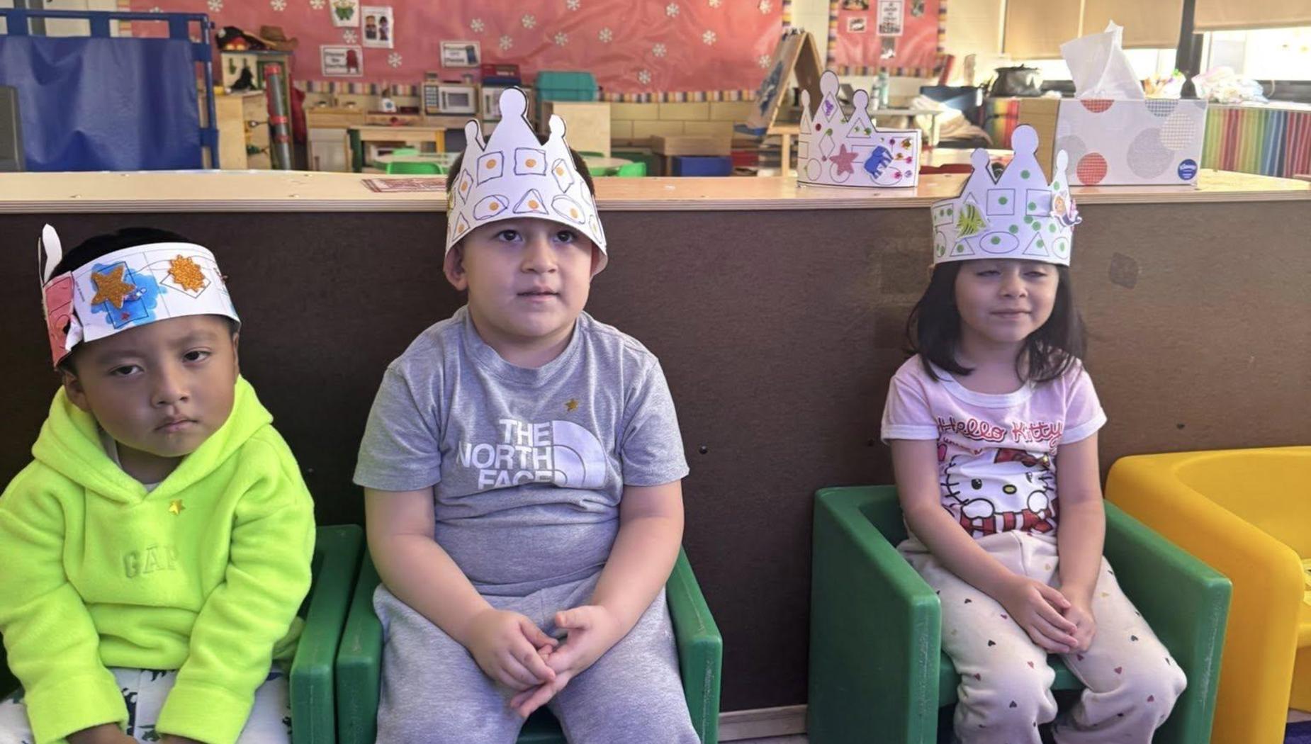 Three children wearing paper crowns sitting on colorful chairs in a classroom.