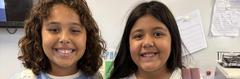 Two smiling children with curly and straight hair standing side by side in a classroom.