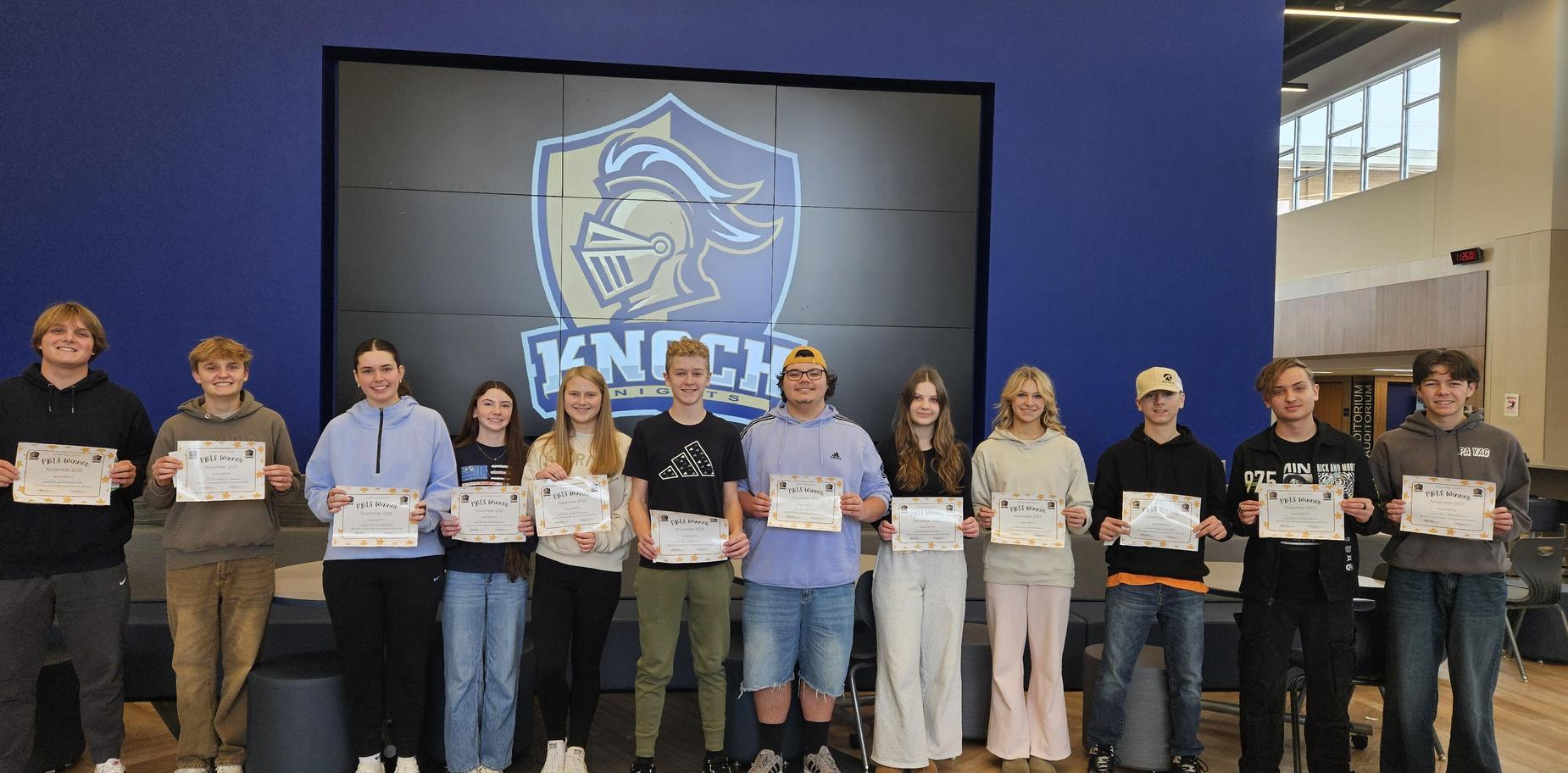 Group of students holding certificates in front of a school logo backdrop.