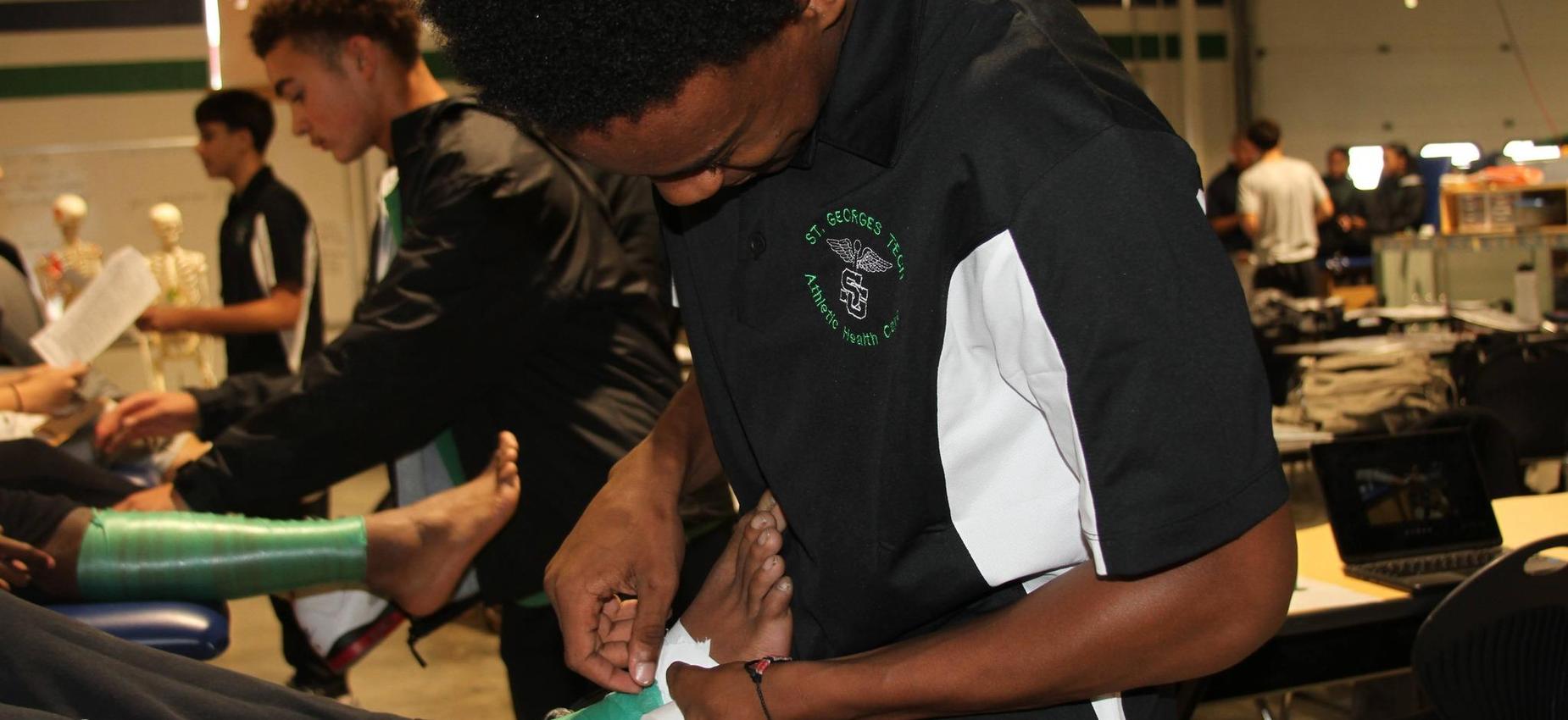 A student wrapping an ankle with bandage, in a classroom setup.