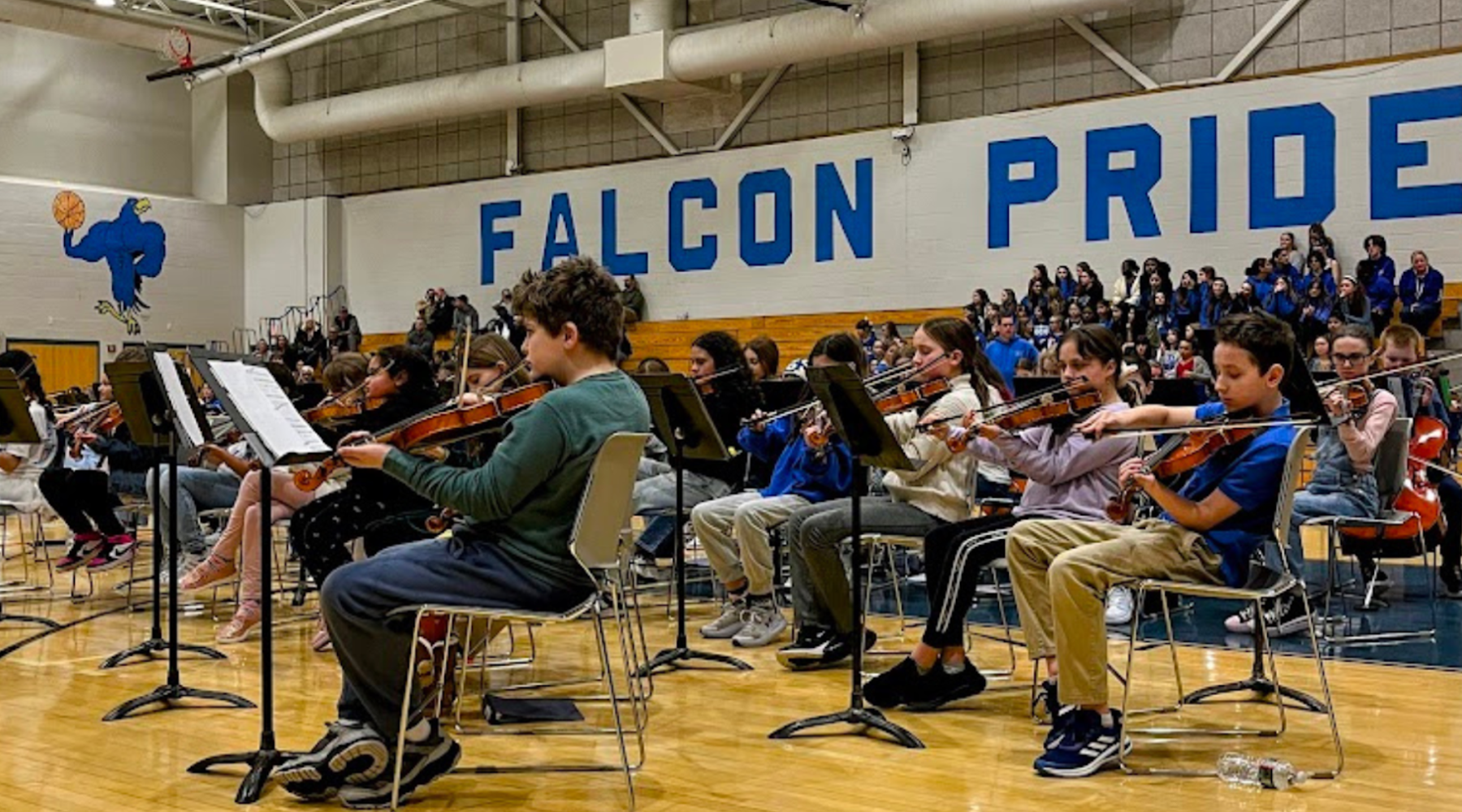 Children playing instruments in a school gym decorated with blue 'Falcon Pride' signage.