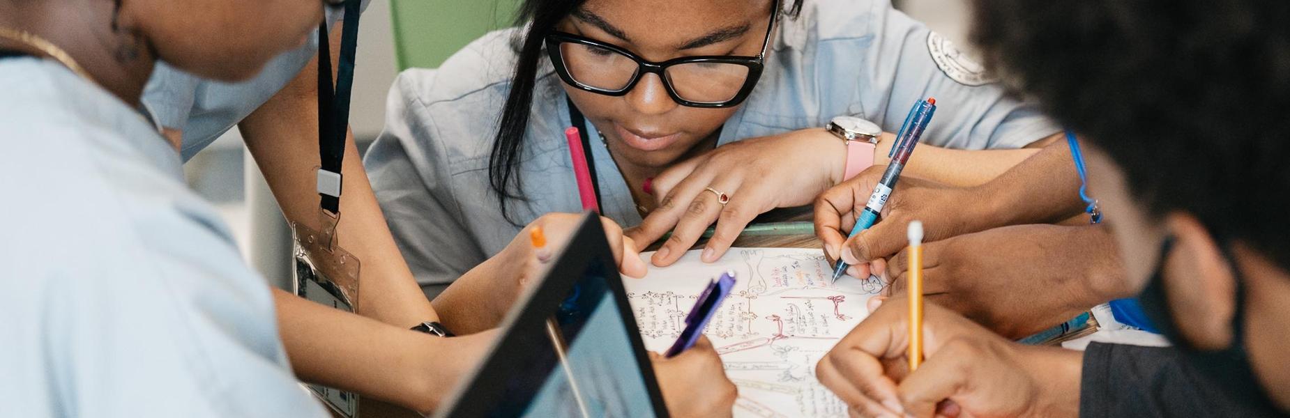 A group of students in gray scrubs are gathered closely around a desk, collaborating on a written assignment. Several hands are visible holding pens and writing in a notebook filled with notes and diagrams. A laptop is partially visible in the foreground, suggesting the use of technology alongside handwritten work. The setting appears to be a classroom with green chairs and a focus on teamwork and active engagement.
