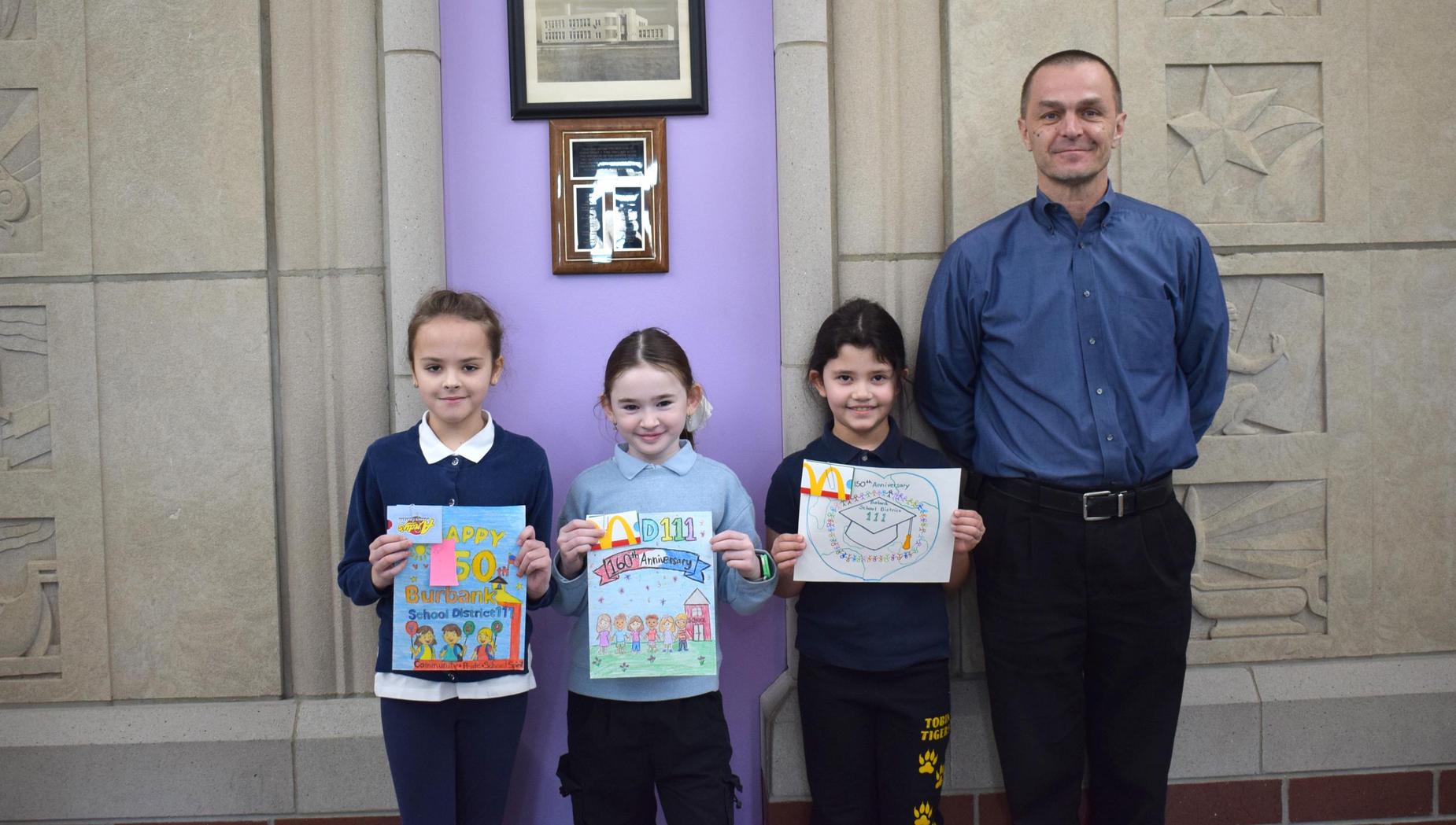Three children and an adult pose with art projects in a school setting.