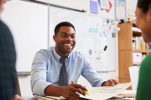 Smiling man in a blue shirt with a tie, discussing paperwork in a classroom.