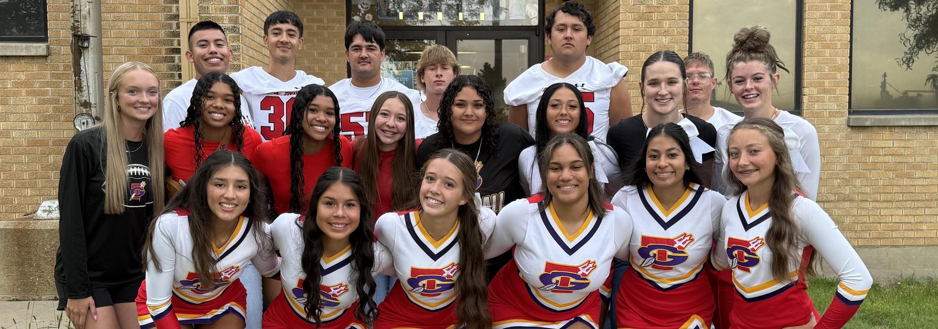 A group of cheerleaders and student athletes posing together outside a school building.