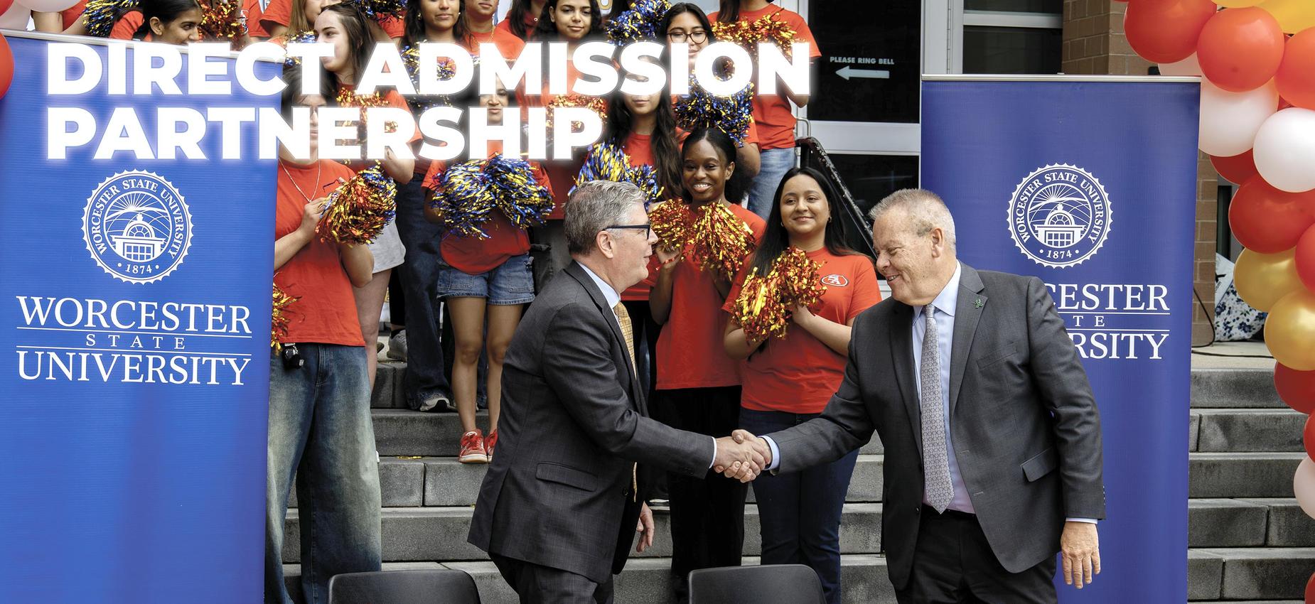 Two men shake hands at an event with a cheering crowd behind them.