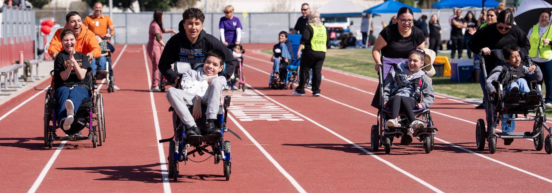 Students using wheelchairs participate in a race on a school track while classmates, staff, and volunteers assist and cheer them on.
