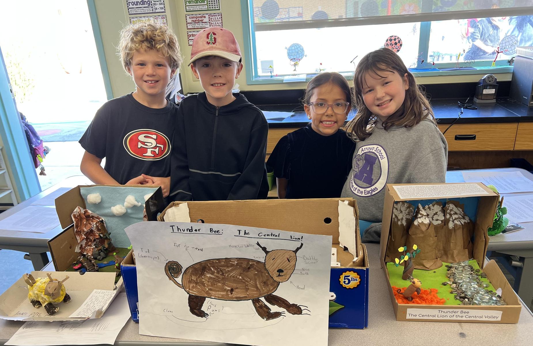 Four children stand together showcasing their projects in front of a table with creative displays.