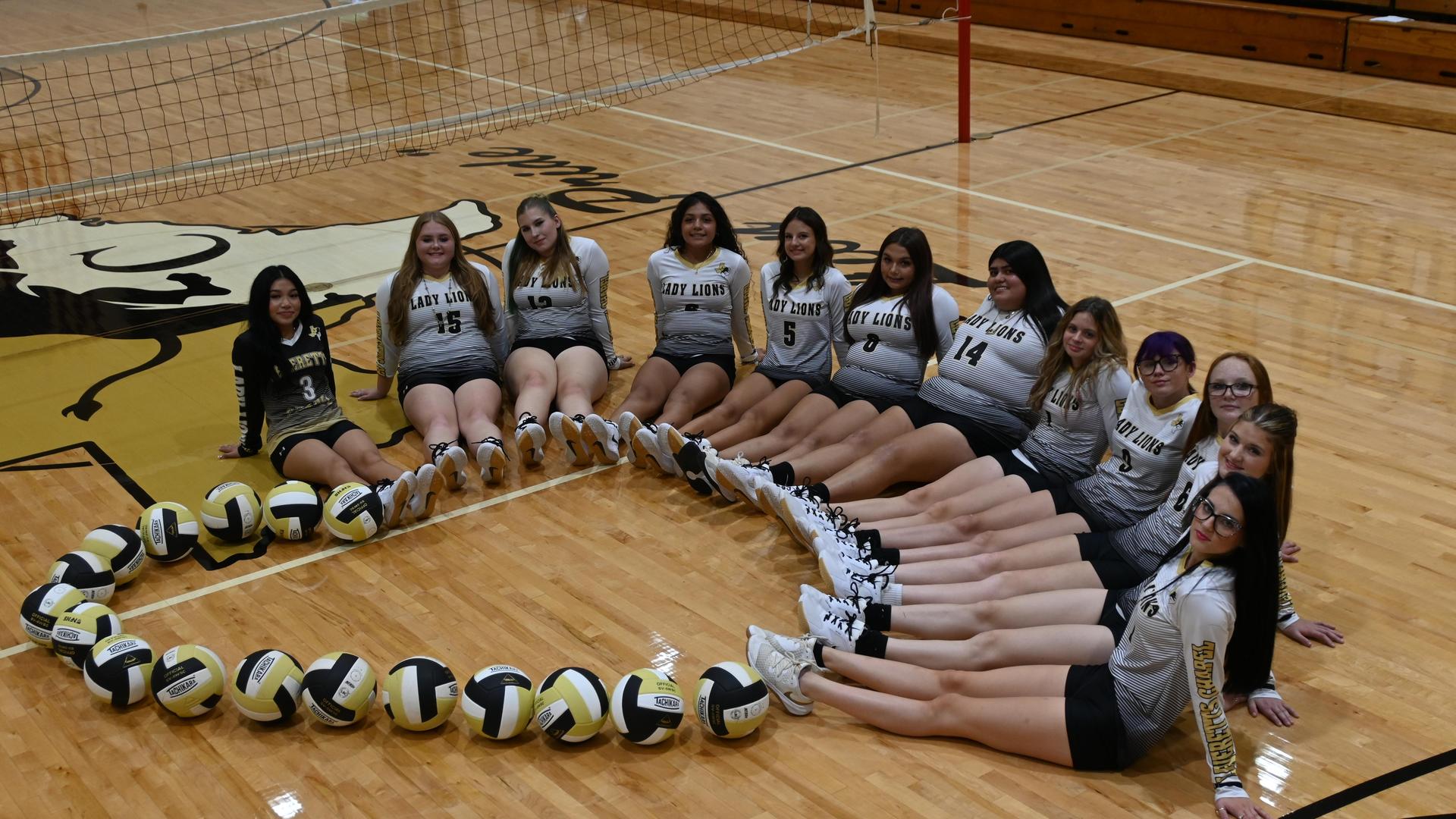 Volleyball team arranged in a circle with volleyballs on a gym floor.