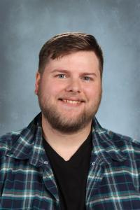 Man with a plaid shirt and V-neck t-shirt smiling against a blue backdrop.