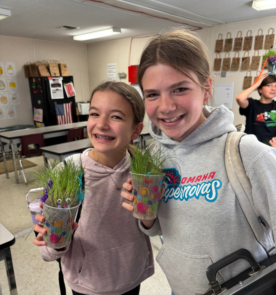 Two girls proudly holding decorated cups of grass in a classroom.