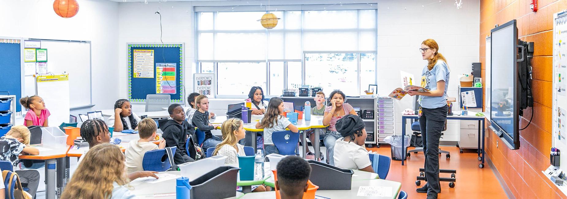 Students listening attentively as a teacher reads in a colorful classroom.