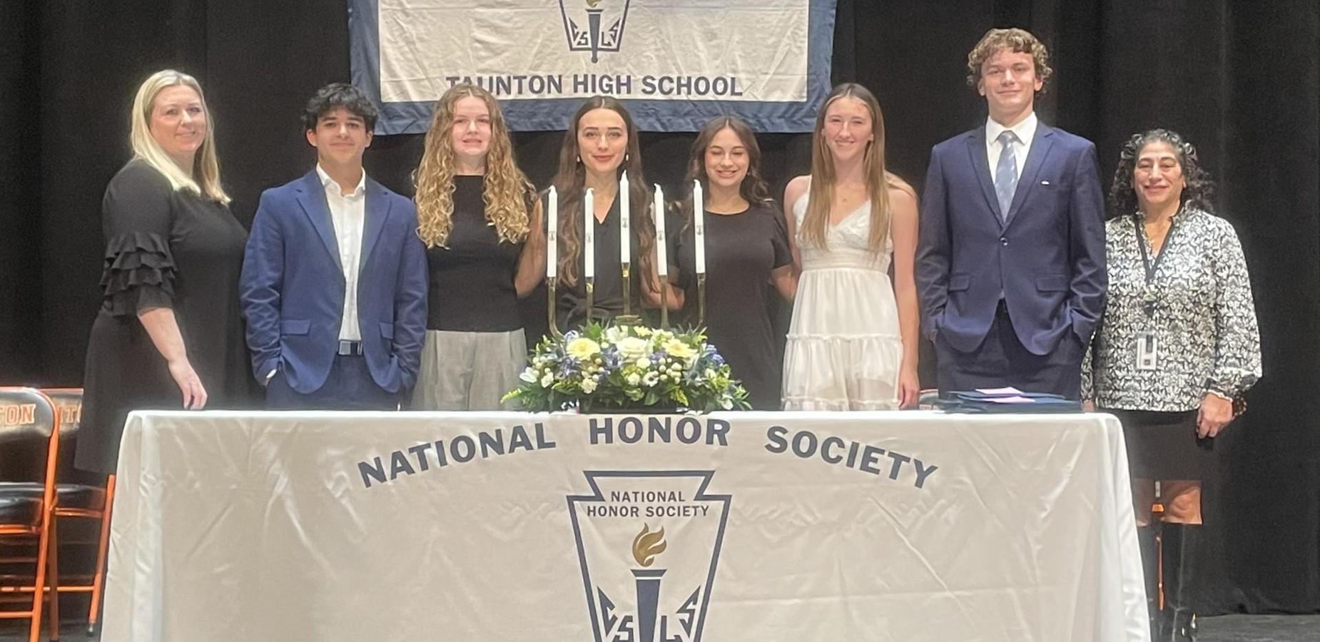 A group of eight students and two adults stand on a stage behind a table draped with a National Honor Society cloth. A National Honor Society banner hangs behind them. The students are dressed in formal attire and are standing in a line, smiling. A floral arrangement and ceremonial candles are displayed on the table in front of them.