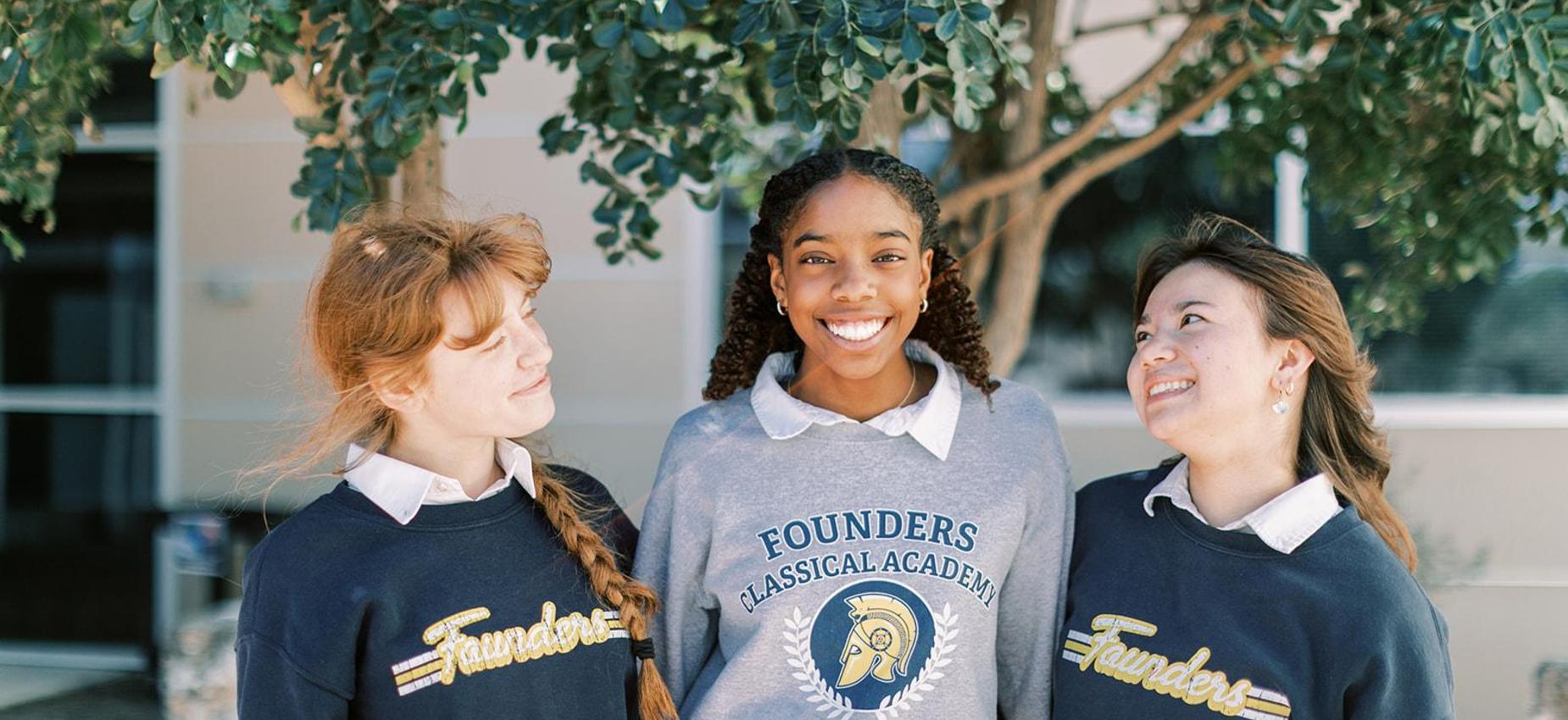 Three smiling students in Hoplites sweatshirts stand together outside
