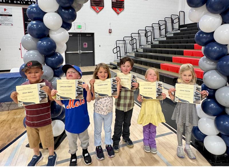 Six smiling elementary school students stand under a balloon arch holding awards certificates