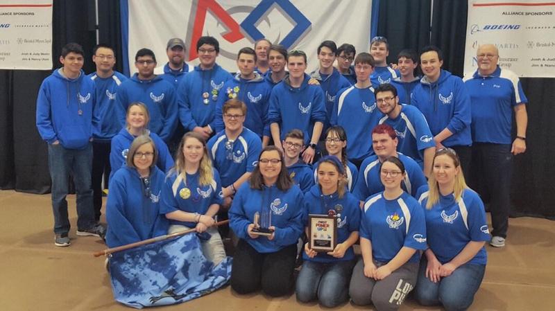 High School Robotics Team dressed in blue shirts holding their awards, a plaque and a crystal award.