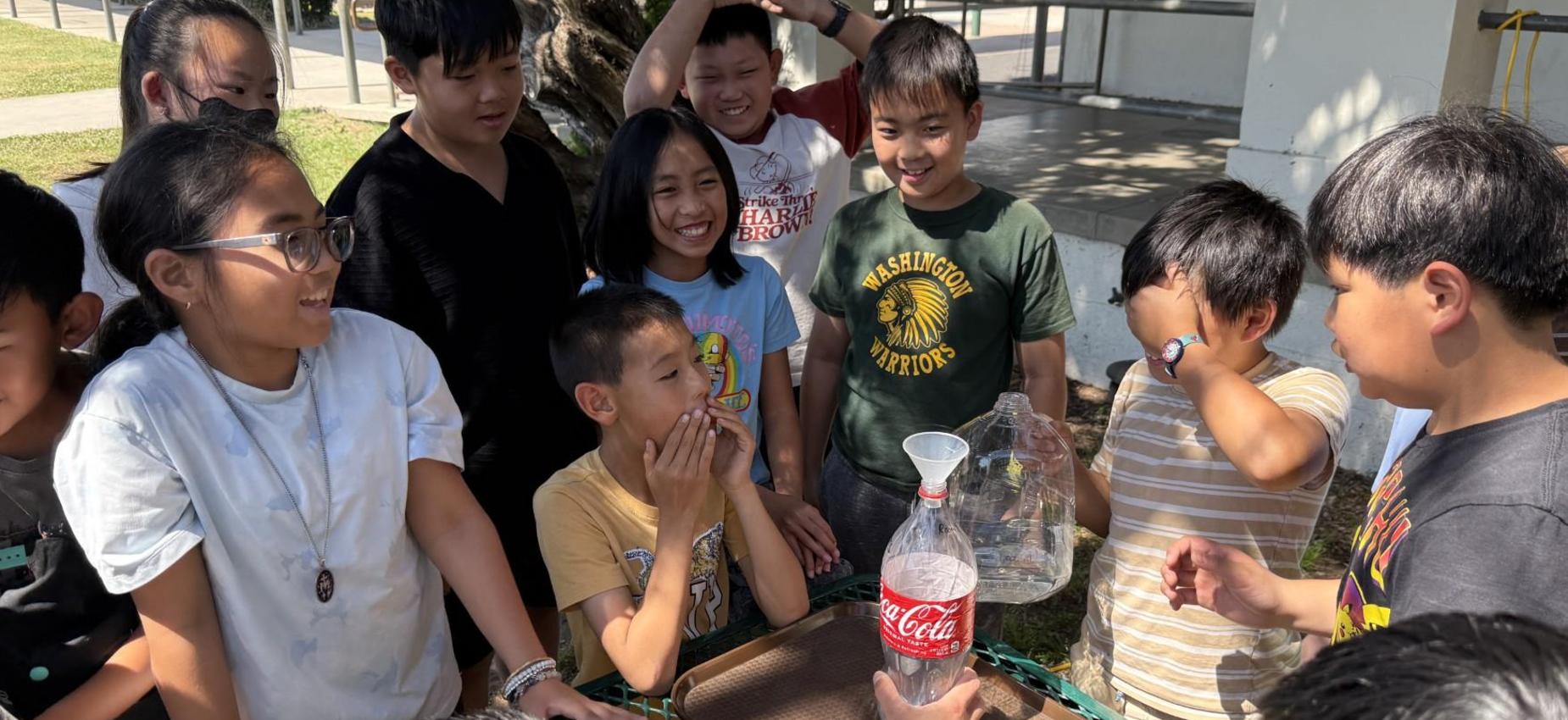 A group of excited children gathered around, observing a science experiment with bottles.