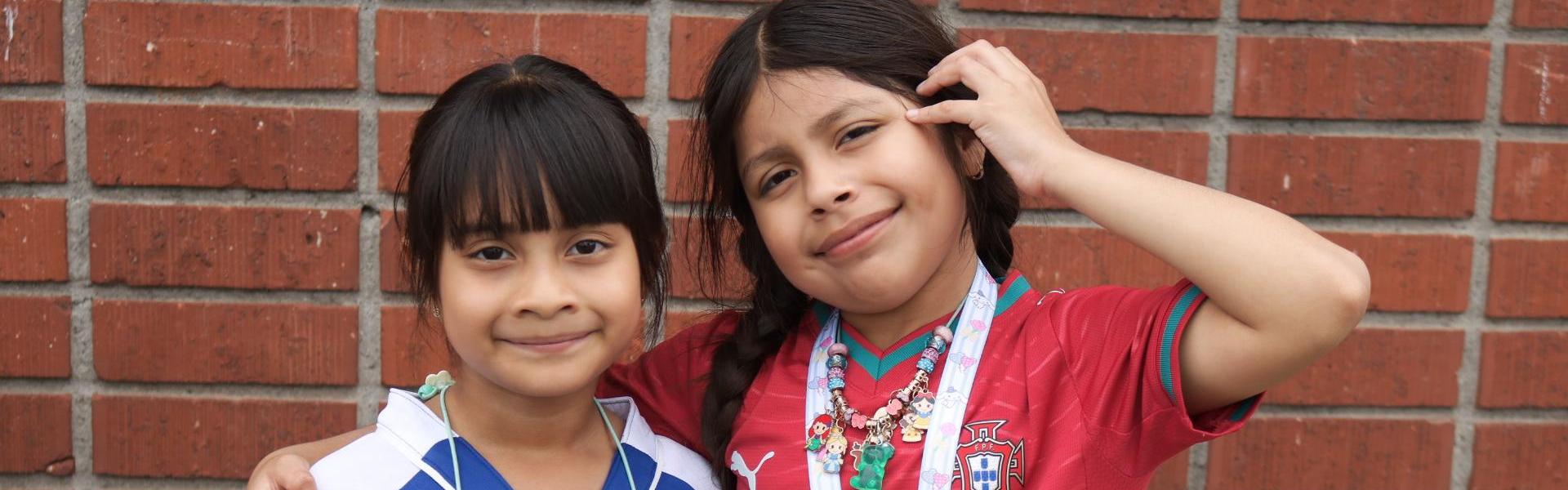 Two girls pose together, one in a blue jersey and the other in a red jersey.