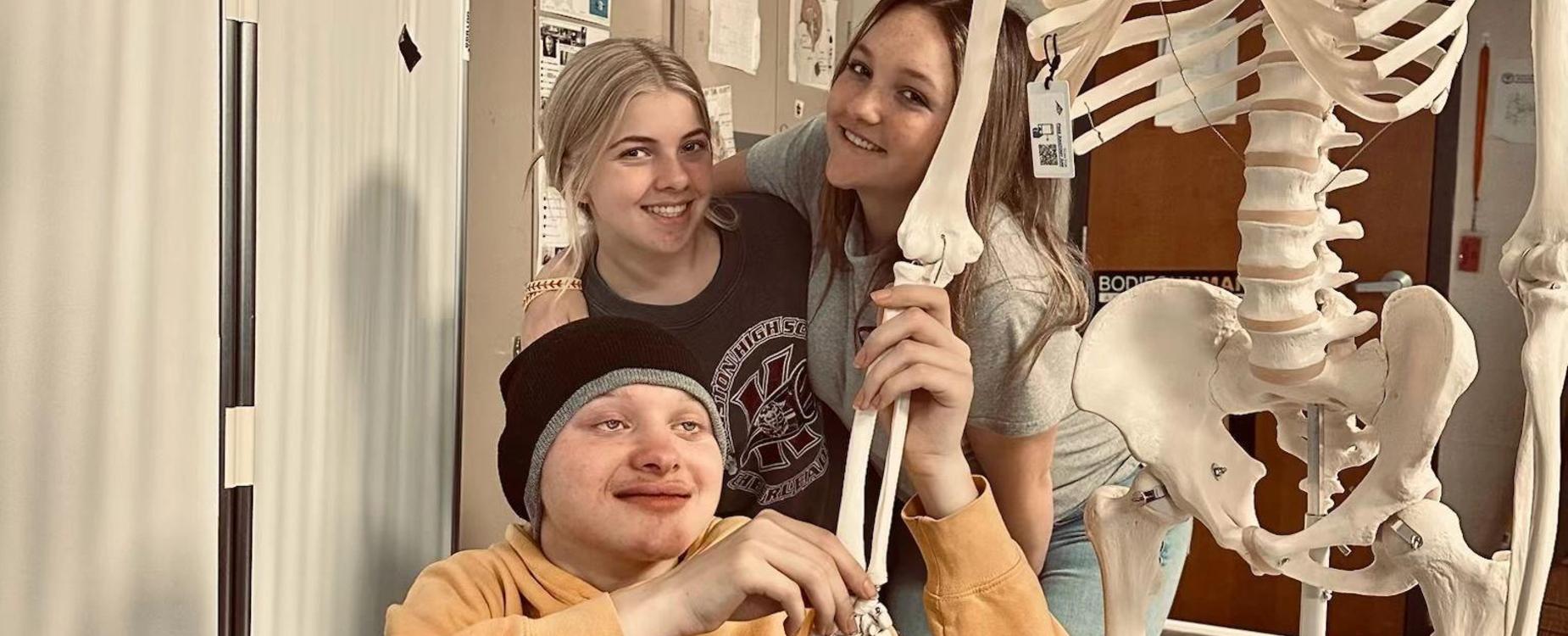 Three students posing together with a skeleton model in a classroom setting.