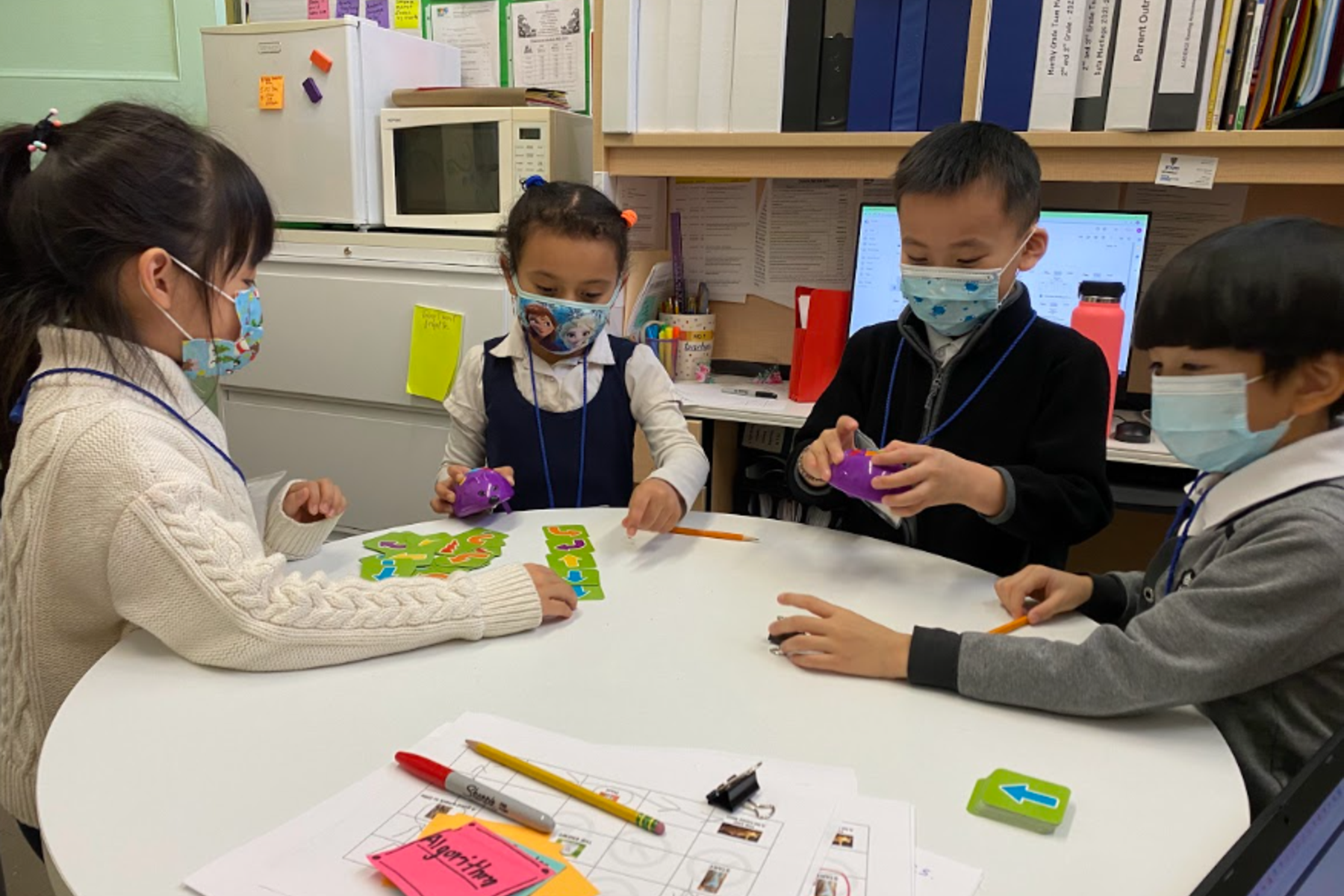 Four children, wearing masks, collaborate at a table with educational materials and crafts.