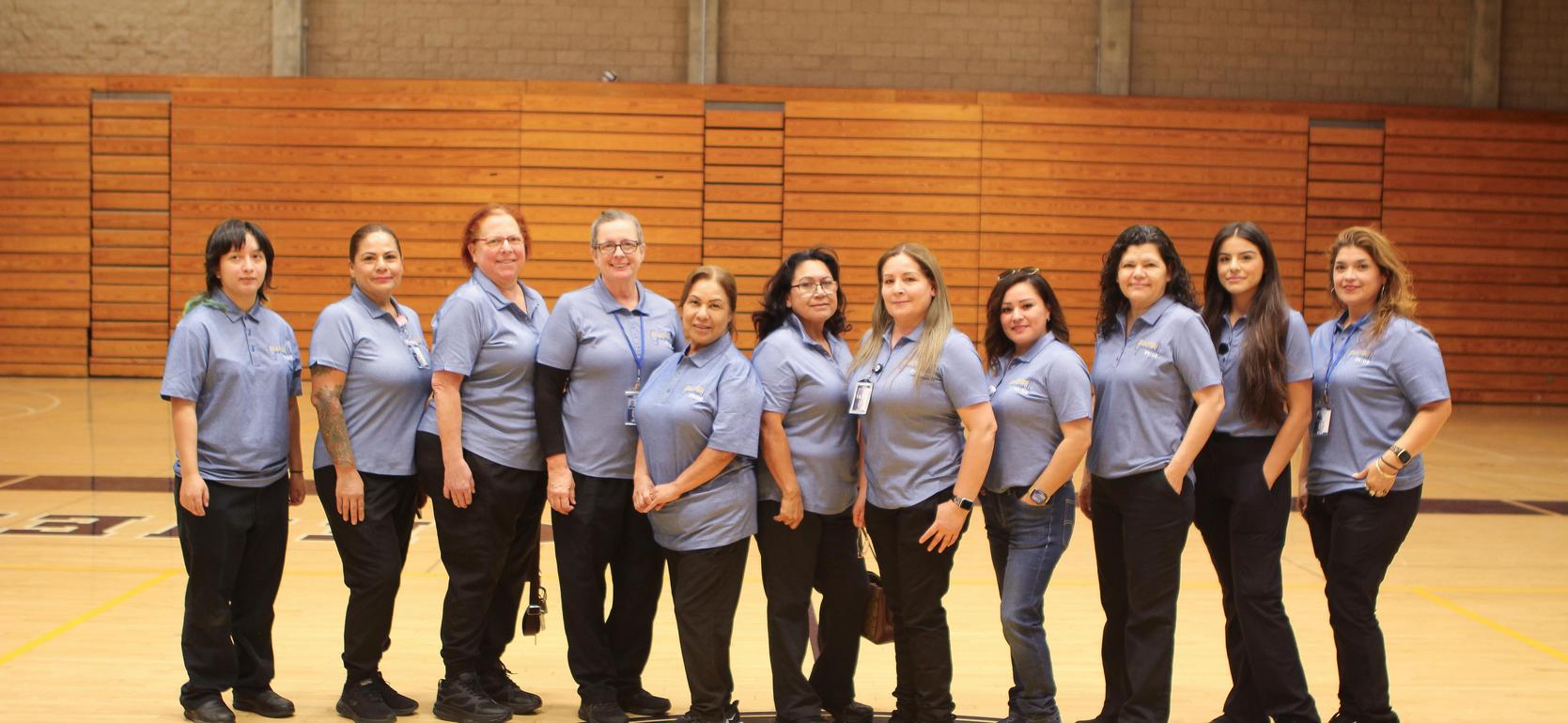Twelve women in matching blue shirts stand together on a gym court with wooden floors.