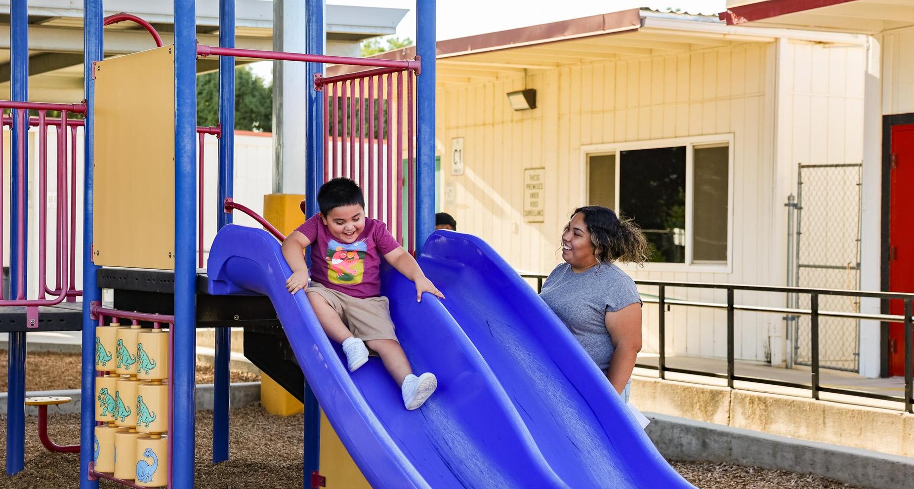 Student happily sliding down slide while parent watches