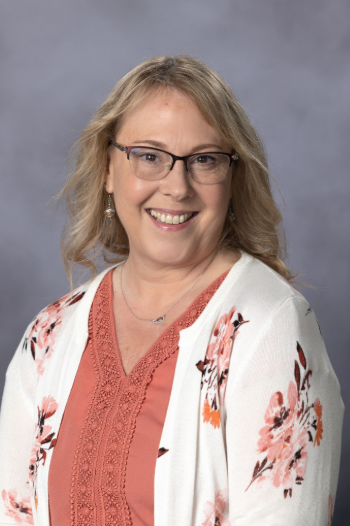 Smiling woman with glasses and a floral cardigan against a gray background.