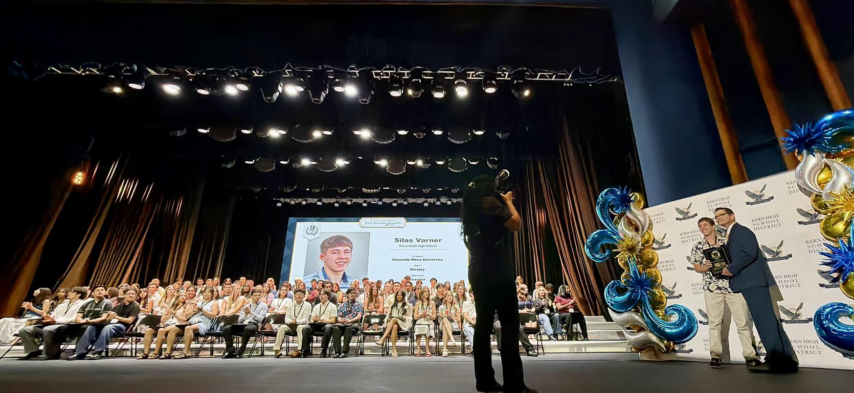 A stage with a crowd seated, a presenter with a trophy, and a large screen displaying a portrait.