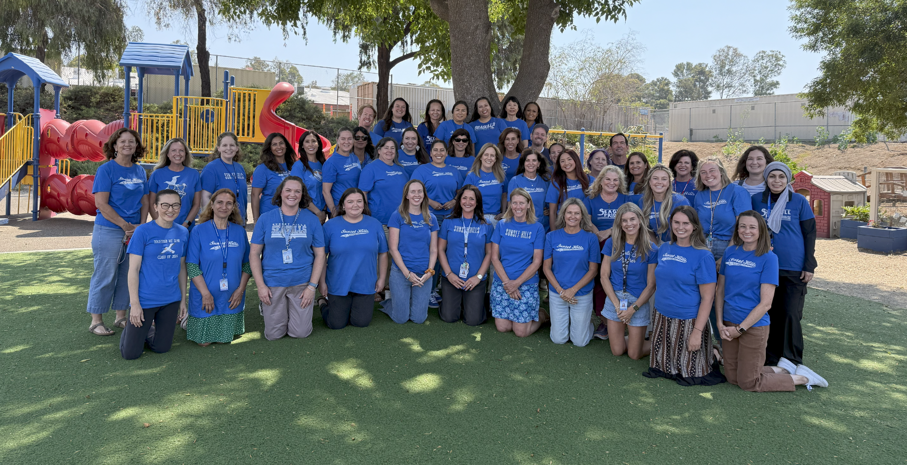 photo of staff wearing blue shirts in front of a tree and playground structure.