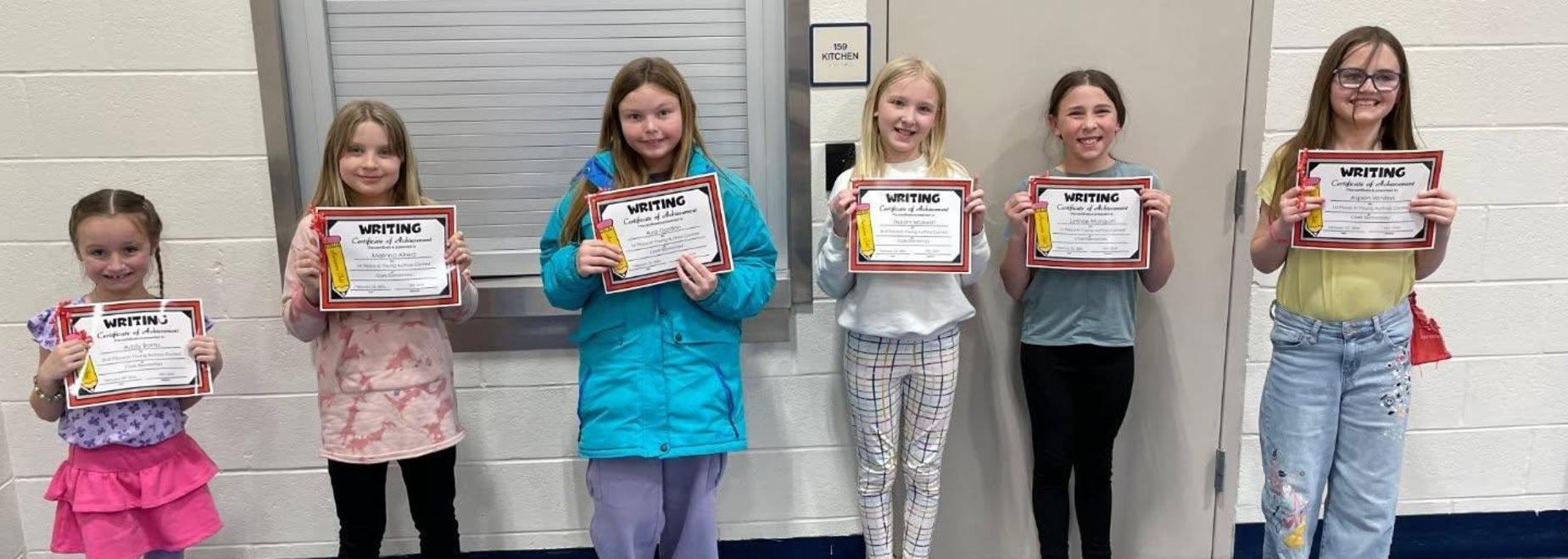 Six children holding writing achievement certificates indoors.