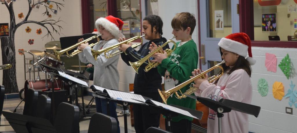 four students play the trumpet, 2 are wearing Santa hats