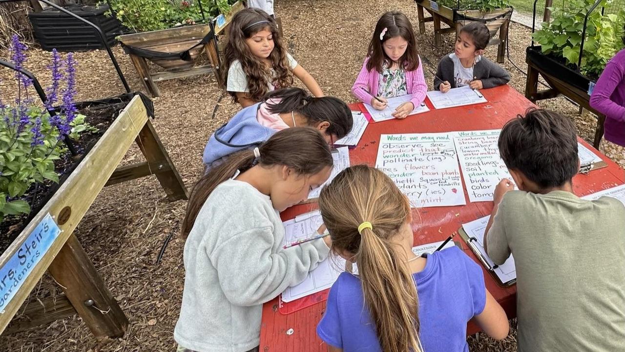 Several children writing and drawing at a colorful outdoor table in a garden.