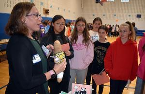 A woman explains an object to a group of attentive girls in a gymnasium.