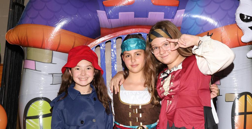 3 Girls wearing Halloween costumes standing in front of a balloon in the shape of a bat.