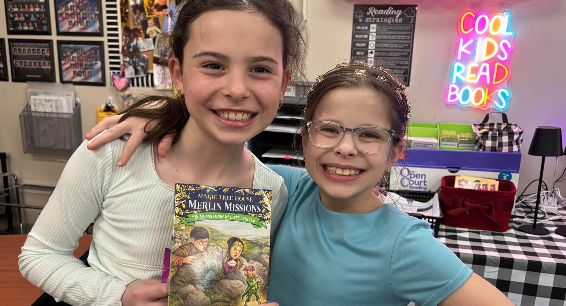 Two students stand together with big smiles and one holds a book