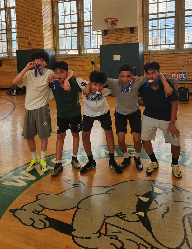 Five boys celebrating together in a gym, wearing medals and posing with arms around each other.
